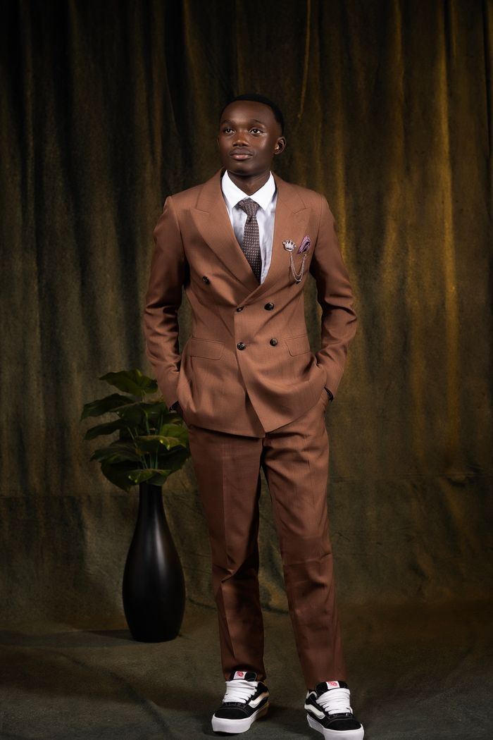 about-us Man in brown suit and sneakers with plant indoors, warm lighting.